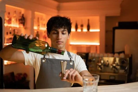 Young bartender in gray apron pours alcoholic drink to steel jigger beaker fr Stock Photos