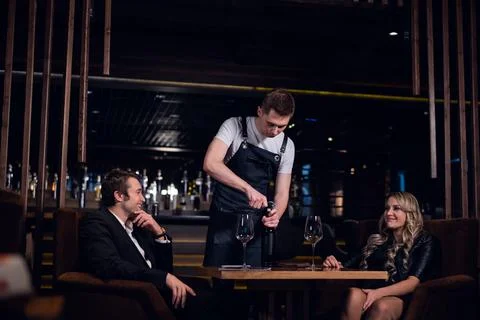 A young bartender opens a bottle of red wine for a man and a woman at a table in Stock Photos