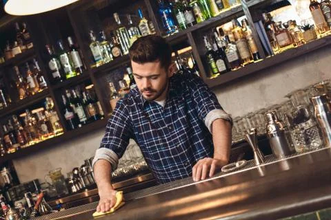 Young bartender standing at bar cleaning counter thoughtful Stock Photos