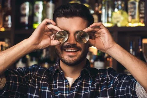 Young bartender standing at bar counter covering eyes with jigger cheerful close Stock Photos