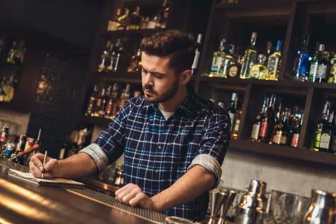 Young bartender standing at bar counter writing receipt concentrated Stock Photos