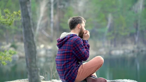Young beard man playing on the harmonica. Sitting on the rock near the lake. Stock Footage 106091250