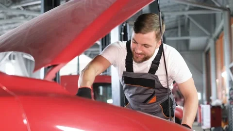 A young bearded auto mechanic inspects a car under the hood in a garage. Stock Footage 212346254