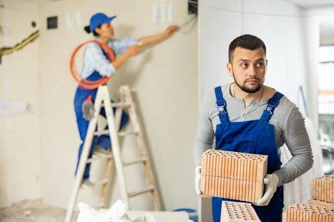 Young bearded builder stacking red bricks on construction site indoors Stockfoto's