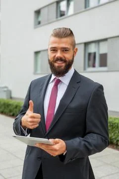 Young bearded businessman using tablet pc outside the office. Thumbs up. Posi Stock Photos