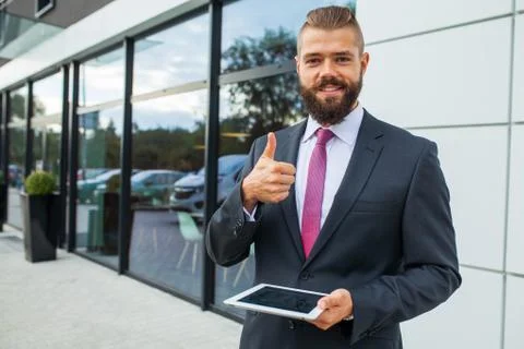 Young bearded businessman using tablet pc outside the office. Thumbs up. Posi Stock Photos