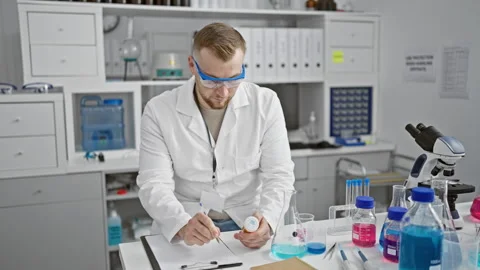 A young bearded chemist in safety goggles and a white lab coat examines a s.. Vidéo 263486261
