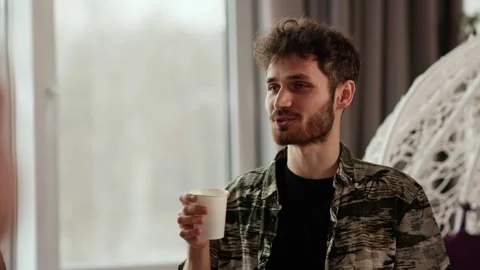 A young bearded guy holds a cup of coffee in his hand and communicates with Stock Footage 243256253