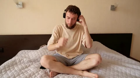 A young bearded guy sitting on a bed with a laptop, takes wireless headphones Stock Footage 279882787