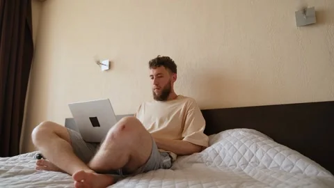 A young bearded guy is sitting with a laptop on his bed, surfing the Internet Stock Footage 279590896