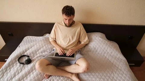 A young bearded guy is sitting with a laptop on his bed, surfing the Internet Stock Footage 279591005