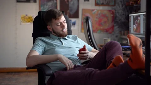 A young bearded guy smokes marijuana while sitting on an armchair. Vidéo 127864409