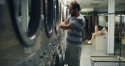 Young bearded guy standing by washing machines and taking out his clothes in int Stock Footage 199456615