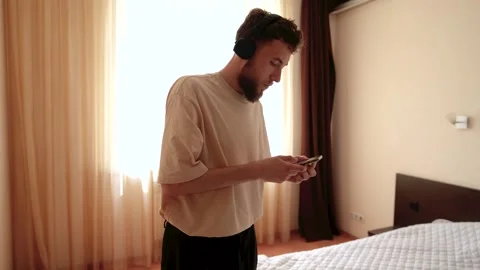 A young bearded guy in wireless headphones selects music on his phone to dance Stock-Footage 278364087