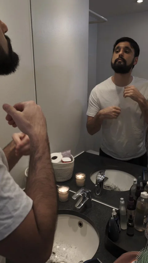 Young bearded man applying beard oil in bathroom mirror, grooming routine Video stock 335637017