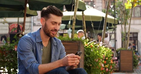 Young Bearded Man browsing his Cellphone and laughing while sitting on terrace Vídeo Stock 111466851
