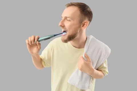 Young bearded man brushing teeth with electric toothbrush on light background Stock Photos