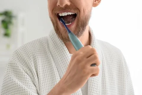 Young bearded man brushing teeth with electric toothbrush in bathroom, closeu Stock Photos