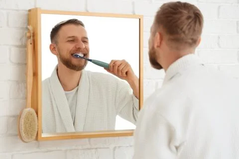 Young bearded man brushing teeth with electric toothbrush near mirror in bath Stock Photos