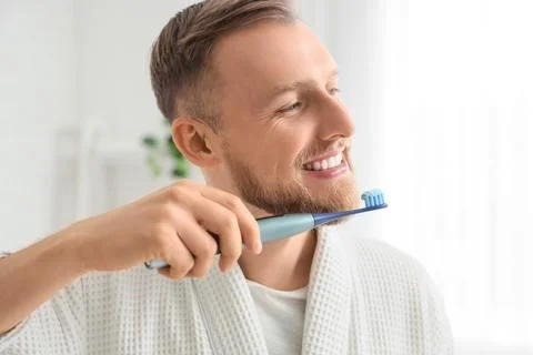 Young bearded man brushing teeth with electric toothbrush in bathroom, closeu Stock Photos
