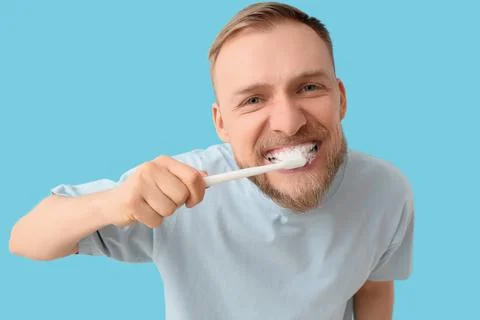 Young bearded man brushing teeth on blue background, closeup Stock Photos