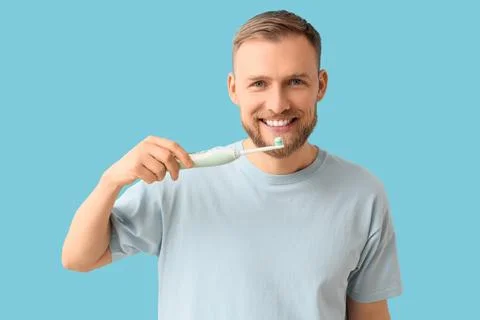Young bearded man brushing teeth with electric toothbrush on blue background Stock Photos