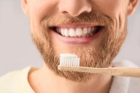 Young bearded man brushing teeth on light background, closeup Stock Photos