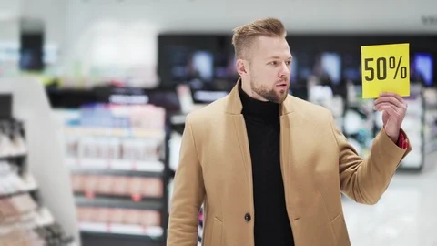 Young bearded man at cosmetics store shows two discount posters with big sale Vidéo 121951419