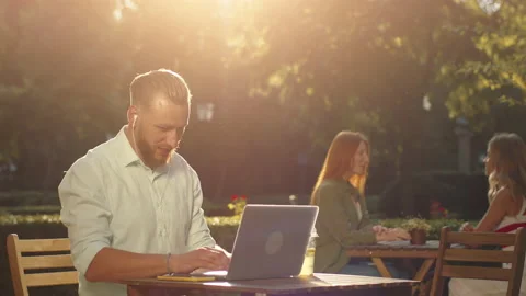 A young bearded man is doing some work on his laptop on a table in the park, in Stock Footage 159712674