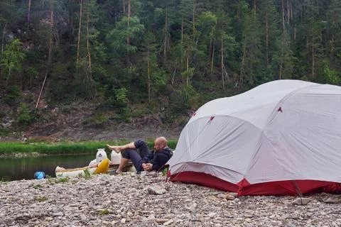 A young, bearded man drinks tea on a camping trip, in nature, against the Stock Photos