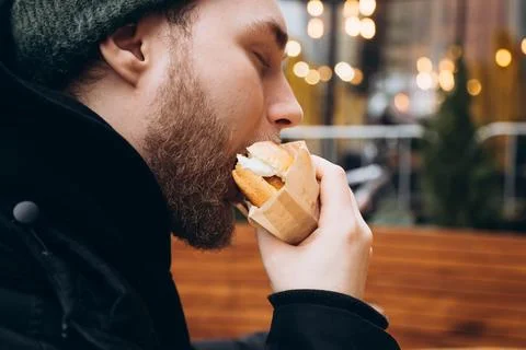 A young bearded man eats a burger outside against the backdrop of a cafe. Stock Photos