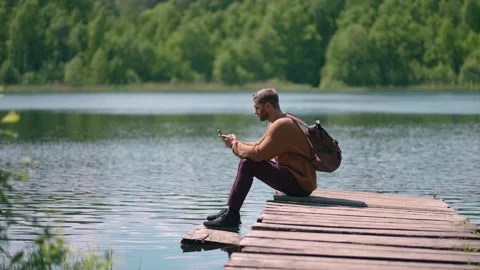 A young bearded man with glasses and a backpack sits on a wooden lake pier in Stock Footage 155400964