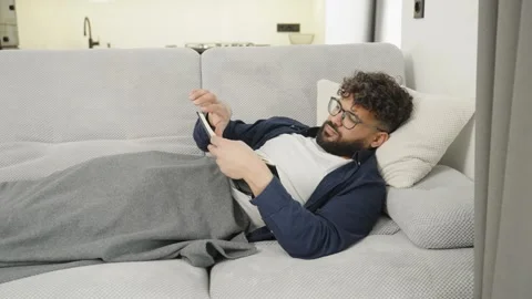 Young bearded man with glasses reading a book on the couch at home Stock Footage 345128407