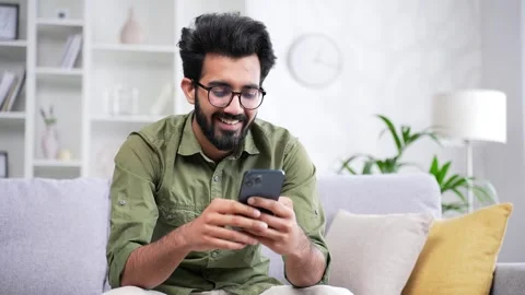Young bearded man in glasses using smartphone sitting on sofa in living room  Stock Footage 252200484