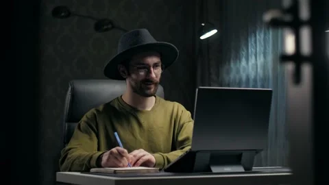 Young Bearded Man learning online using laptop sitting at desk in dark room Stock Footage 232134544