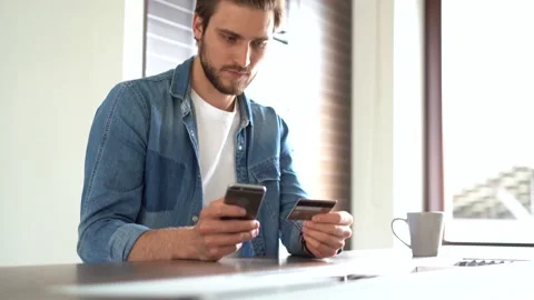 Young Bearded Man makes Online shopping, holding his Credit Card and Smartphone Stock Footage 155332354