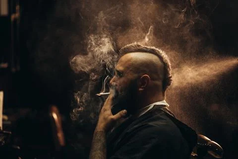 Young bearded man with a mohawk sitting at barbershop and smoking a cigarette Stock Photos