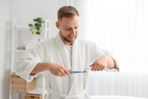 Young bearded man pouring paste onto electric toothbrush in bathroom Stock Photos
