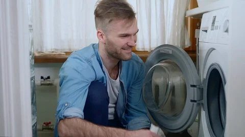 Young bearded man puts clothes in washing machine while sitting on floor in home Stock Footage 130141104