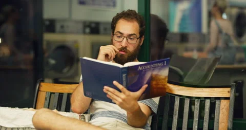 Young bearded man reading a college book on a bench in interior small laundromat Stock Footage 199459455
