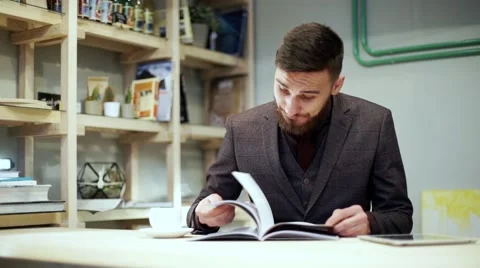 Young bearded man is reading magazine in cafe, drink coffee Stock Footage 56892001