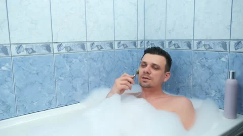Young bearded man is relaxing lying in bath and doing face massage using roller. Stock-Footage 153576277