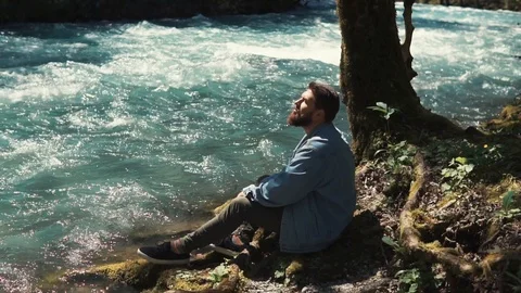 A young bearded man sat down beside a lonely tree and a river to rest Stock Footage 90040559