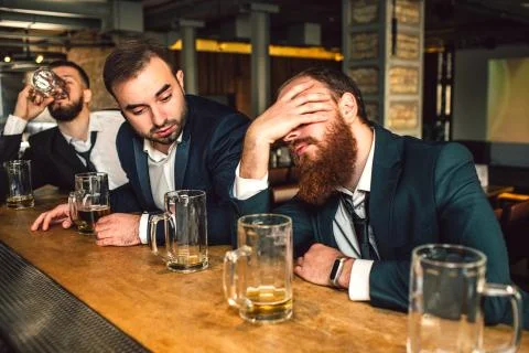 Young bearded man sit in front and sleep. He is drunk. Second young man look at 库存照片