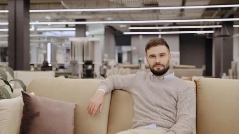 Young bearded man sitting relaxed on a beige sofa in a modern furniture showroom Stock Footage 334654225