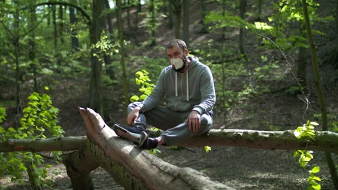 Young Bearded Man Sitting on Tree Branch, Taking Off Face Mask and Checking Stock Footage 130762745
