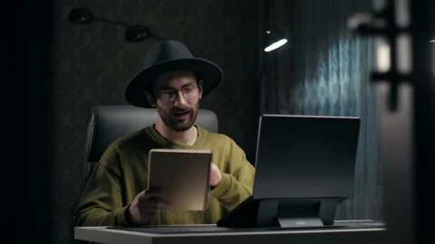 Young Bearded Man teaching online using laptop sitting at desk in dark room Stock Footage 232132326