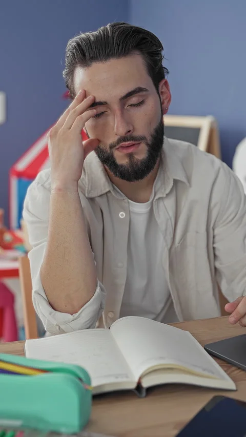 Young bearded man touching forehead with hand, seated at open book and lapt.. Stock Footage 337371231
