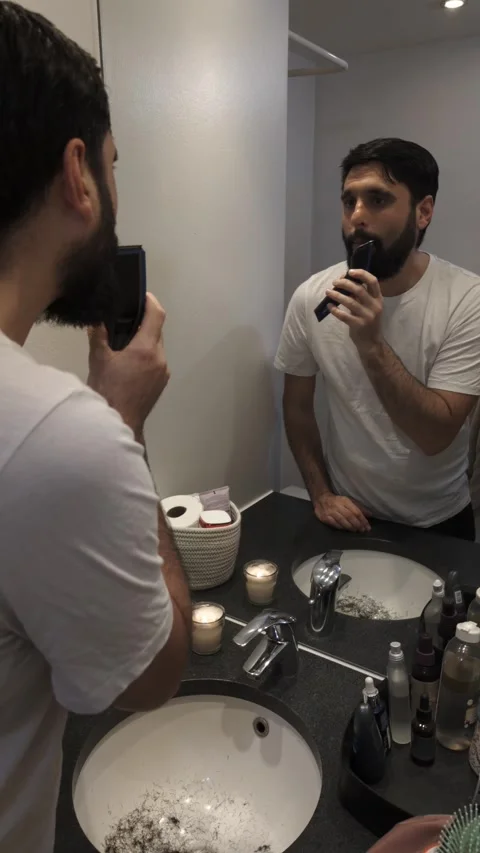 Young bearded man trimming long beard with electric razor in bathroom Video stock 335637566