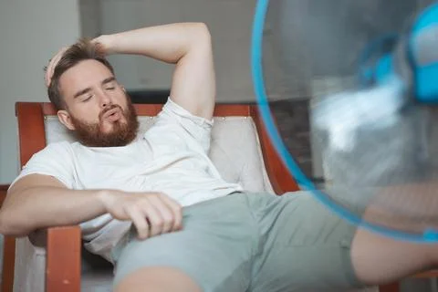 Young bearded man using electric fan at home, sitting on couch cooling off 写真素材